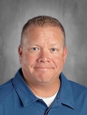 A man with short hair and a friendly expression, wearing a blue shirt, poses for a school portrait against a gray background.
