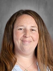 Smiling woman with long brown hair and freckles, wearing a light blue top, against a gray background.