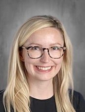 A smiling woman with long blonde hair and glasses poses against a gray background.