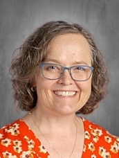 Smiling woman with short curly hair, wearing glasses and an orange floral shirt, set against a gray background.