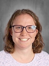 Smiling woman with curly hair and glasses, wearing a light purple sweater, against a gray background.