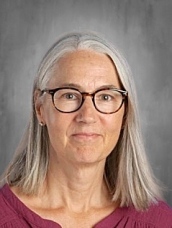 A woman with long gray hair and glasses smiles, wearing a maroon top against a neutral background.