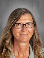 A smiling woman with long, wavy hair and glasses, wearing a light gray top, against a plain background.