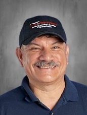 A smiling man with a mustache wears a black cap and a blue polo shirt, set against a neutral background.