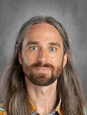 Portrait of a man with long hair and a beard, wearing a patterned shirt, against a neutral background.