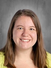 Smiling woman with long brown hair, wearing a bright yellow shirt, against a neutral gray background.