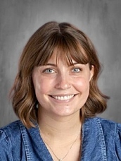 Smiling woman with shoulder-length brown hair, wearing a denim top, against a gray background.