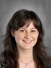 A smiling person with long, dark hair and a necklace, wearing a black and white patterned top against a gray background.