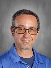 A man with glasses and short gray hair smiles in a blue shirt against a neutral background.