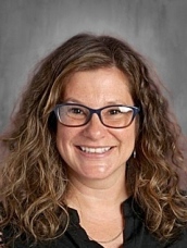 Smiling woman with curly brown hair and glasses, wearing a black top, against a neutral gray background.