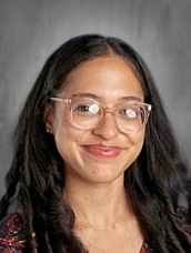 A smiling individual with long, dark hair and glasses, wearing a patterned top against a gray background.