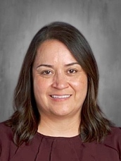 A woman with long dark hair, wearing a maroon top, smiling against a gray background.